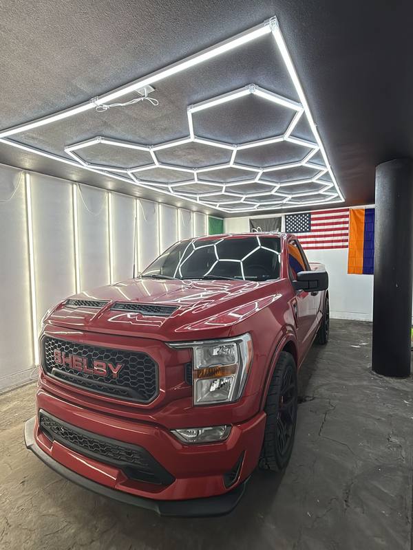 Shelby F-150 in deep candy red under the hexagon LED array, the array mirrored across the hood and windshield; blacked-out SHELBY grille, black wheels, in the restyling bay