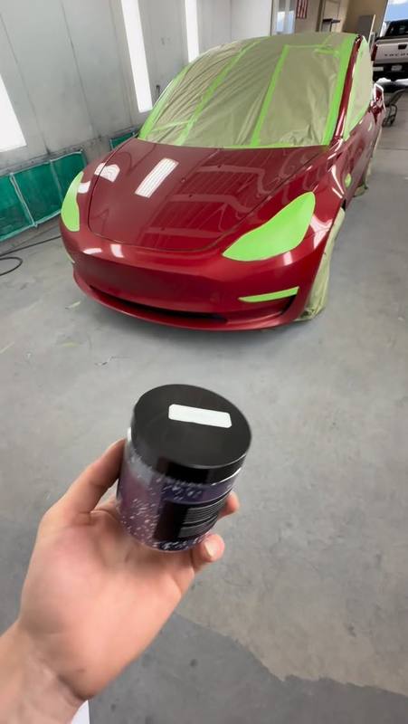 Red Tesla Model 3 masked up in the booth before a Peel Clear color change, with green tape along the headlight, grille and panel edges and paper over the glass and roof, and a Peelclear tin held up in front of it