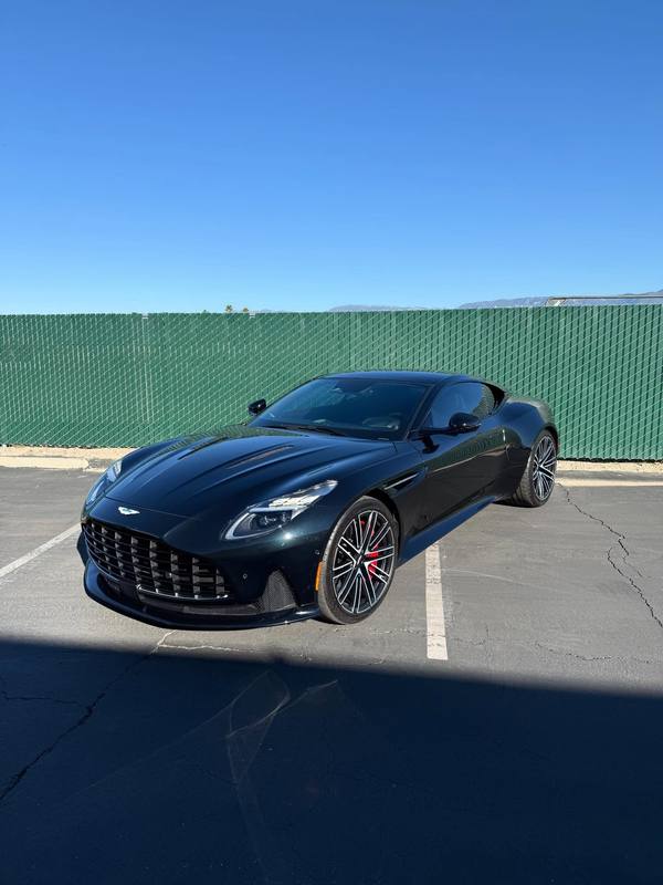 Aston Martin DB12 in dark green parked in the lot outside the shop, the color reading almost black in the shade and turning green across the lit top of the fender and door