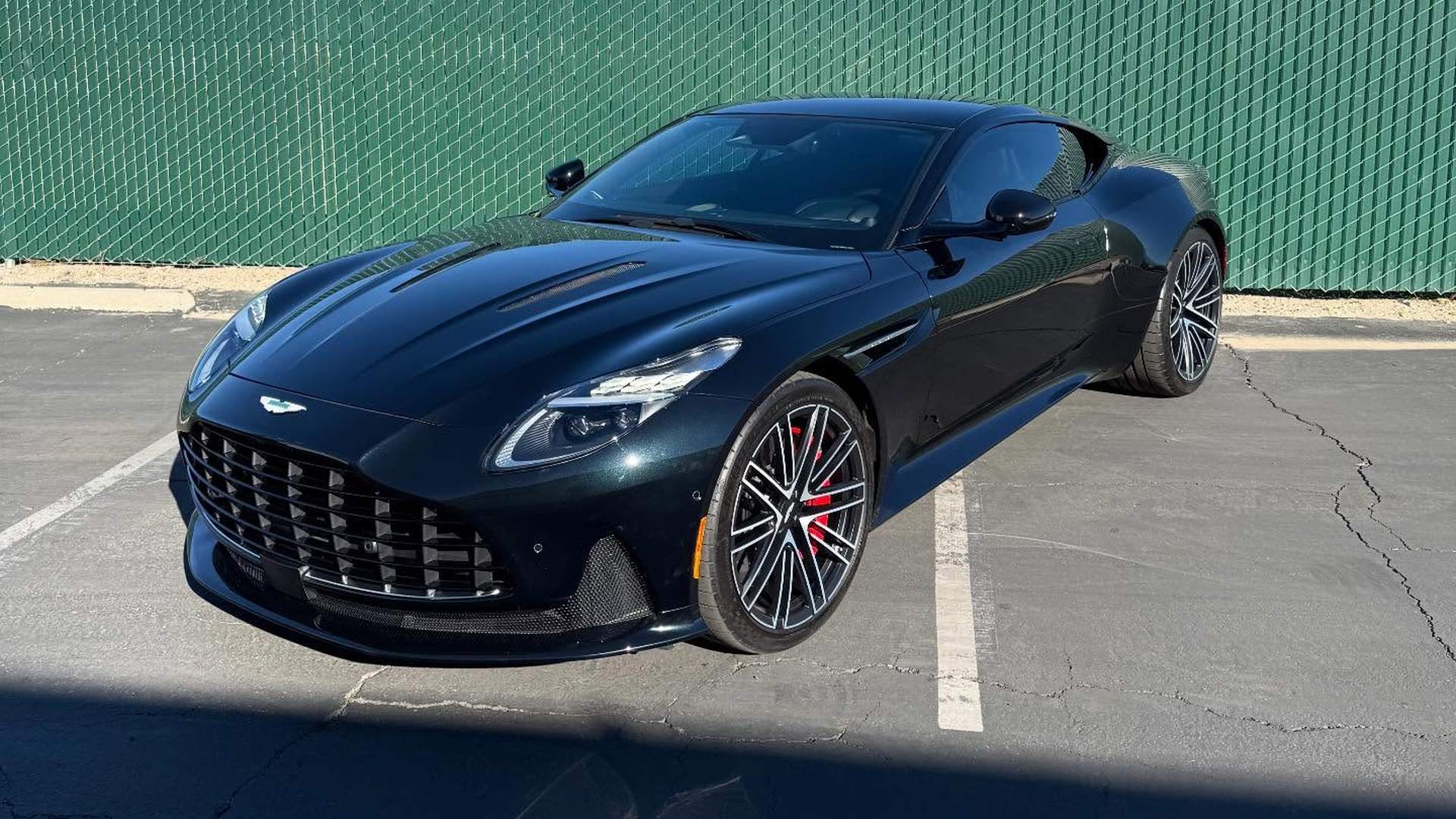Aston Martin DB12 in dark green parked in the lot outside the shop, the color reading almost black in the shade and turning green across the lit top of the fender and door