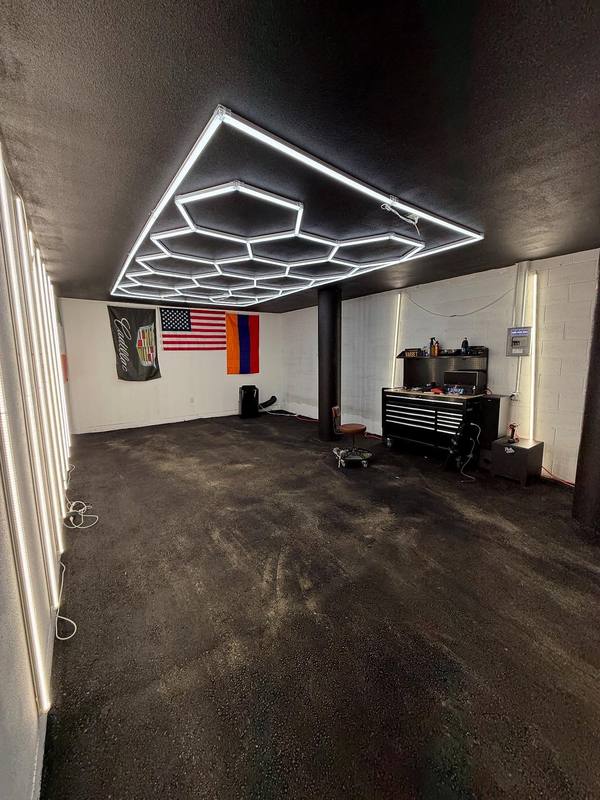 The restyling bay standing empty, a hexagon LED array across the black ceiling, backlit white wall panels down one side, a rolling tool chest against the far wall and three flags hung above it