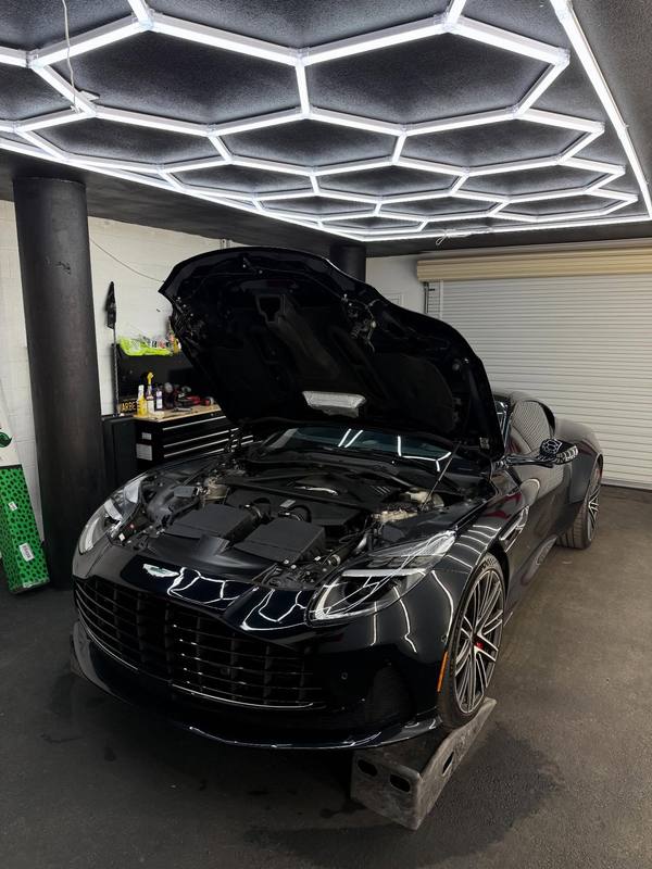Aston Martin DB12 in gloss black with the hood raised under the hexagon LED array, the engine bay open and the array reflected along the fenders and the painted underside of the hood