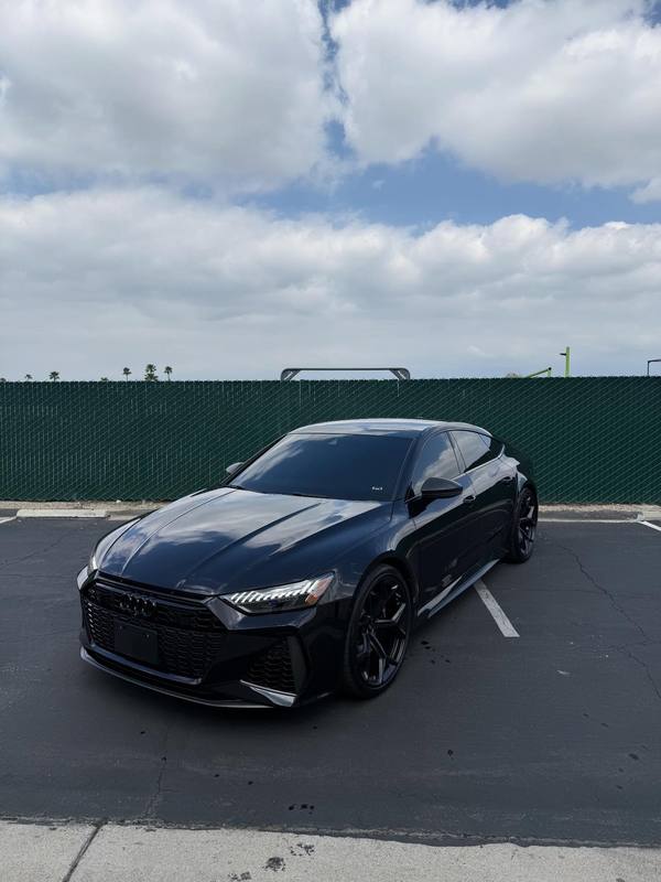 Audi RS7 in gloss black with the rings, grille frame, window surrounds and wheels all black, so the only break in the color is the cloud reflection running down the hood and roof; parked outside the shop