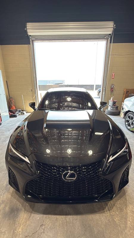 Lexus IS F Sport with ceramic window tint, seen head-on, the side and rear glass reading dark against the bright open roll-up door behind it while the ceiling lights reflect off the black hood