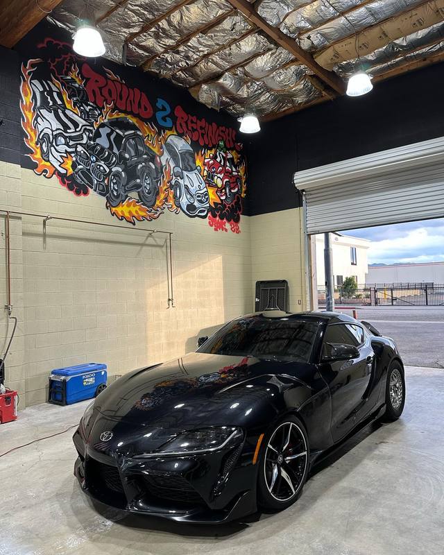 Toyota GR Supra in gloss black inside the shop, the flame mural on the wall behind it reflected across the hood and roof; bronze wheels and dark glass