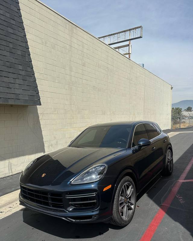 Porsche Cayenne Turbo in dark navy blue parked along the shop's block wall, the gloss deep enough to carry a clean reflection of the wall and sky down the hood, doors and fenders