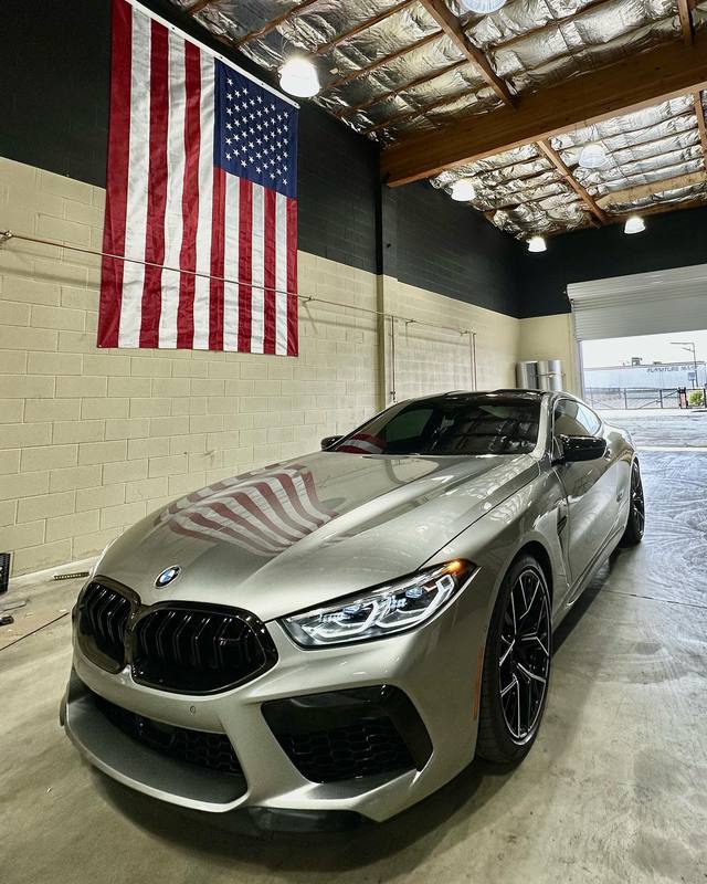 BMW M8 Competition in warm grey parked inside the shop, its hood reflecting the American flag on the wall clearly enough to count the stripes; black kidney grille, black wheels