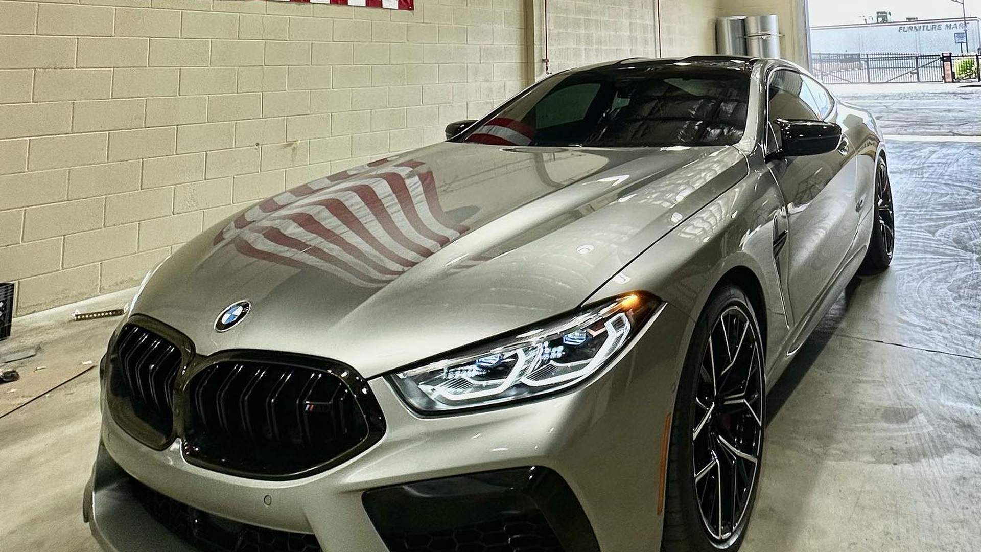 BMW M8 Competition in warm grey parked inside the shop, its hood reflecting the American flag on the wall clearly enough to count the stripes; black kidney grille, black wheels