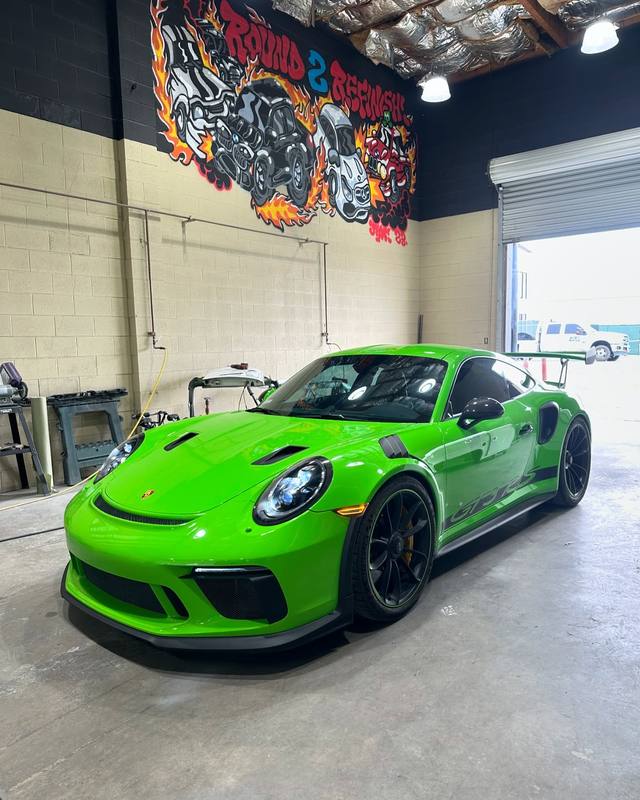 Porsche 911 GT3 RS in bright green inside the shop, rear wing up, black wheels and yellow brake calipers, parked under the flame mural with the roll-up door open behind it