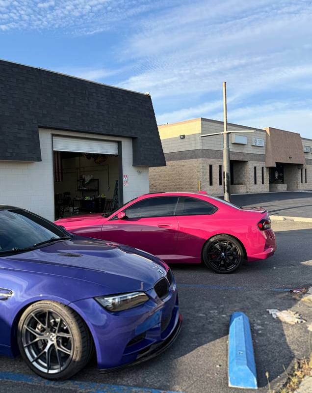 Two color changes side by side outside the shop: a hot pink vinyl-wrapped BMW 2 Series with the pink carried over its bumpers, door handles and mirrors, and a deep purple Peel Clear BMW M3 parked in front of it