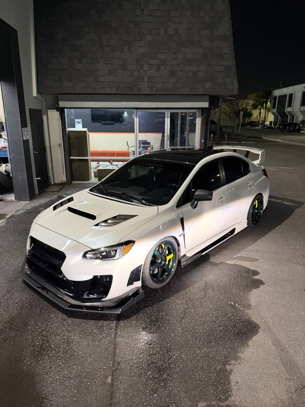 Subaru WRX STI in pearl white with a gloss black roof and black carbon-look splitter, side skirts and wing uprights, photographed outside the shop at night on teal wheels with yellow calipers