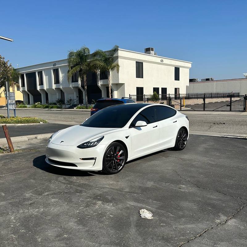Pearl white Tesla Model 3 with dark glass all round, black window surrounds and graphite wheels with red calipers, parked in the lot outside Round 2 Refinish