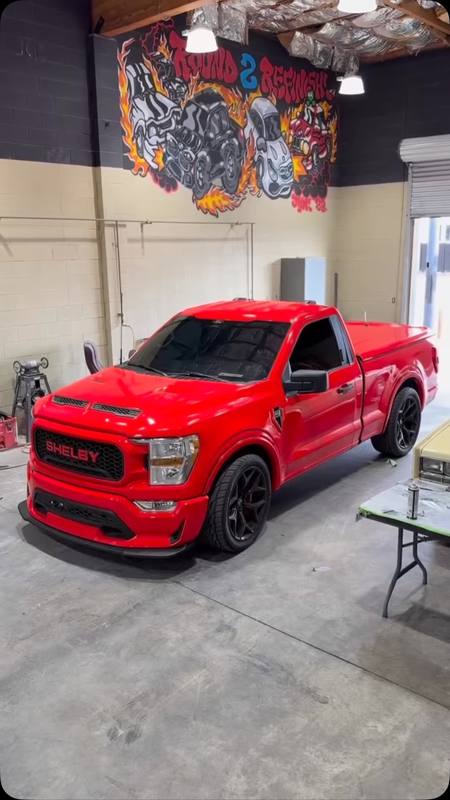 Red Shelby F-150 inside the shop in front of the flame mural, lowered on black wheels with the blacked-out SHELBY grille and the hood vents standing out against the gloss red body