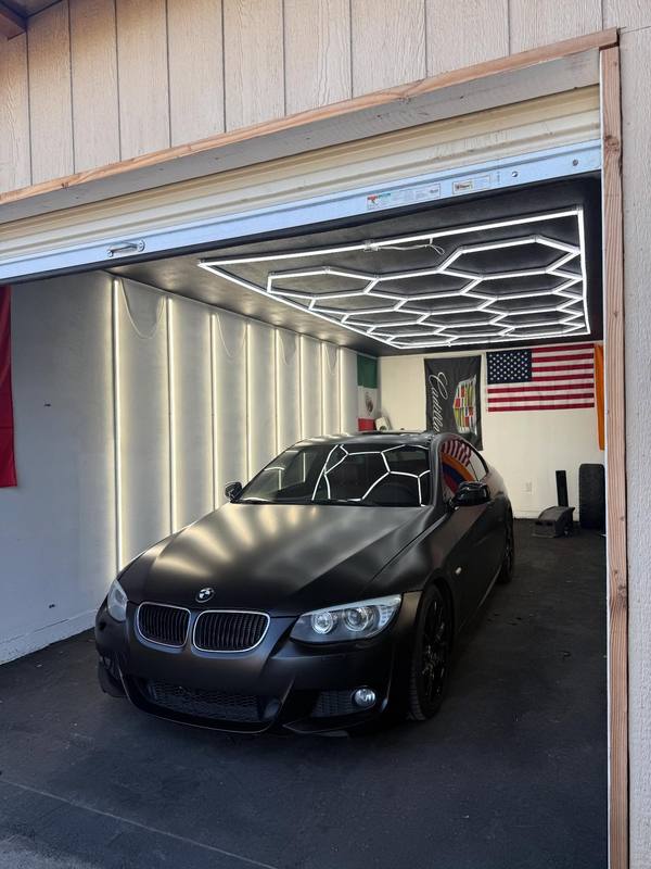 BMW E92 coupe in flat satin black under the hexagon LED array in the restyling bay, the array showing on the hood as a soft glow rather than a hard outline, with the kidney grille surrounds left bright