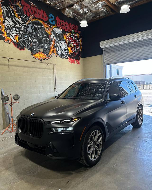 BMW X7 in flat satin black with the grille, badges and window trim all dark, so daylight from the open roll-up door spreads as a soft band across the hood instead of a sharp reflection; inside the shop under the flame mural