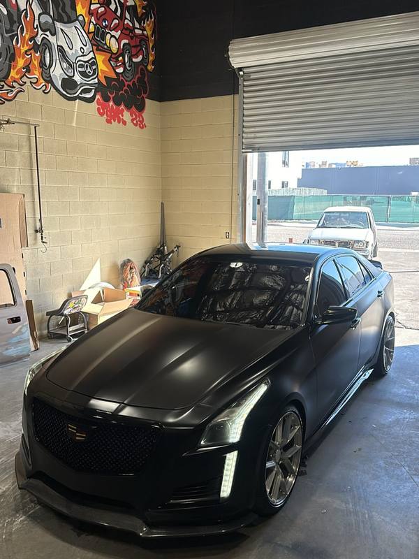 Cadillac CTS-V in flat satin black with a gloss black splitter and side skirt, the matte hood holding only a soft band of daylight from the open shop door and the Cadillac crest left in color on the mesh grille