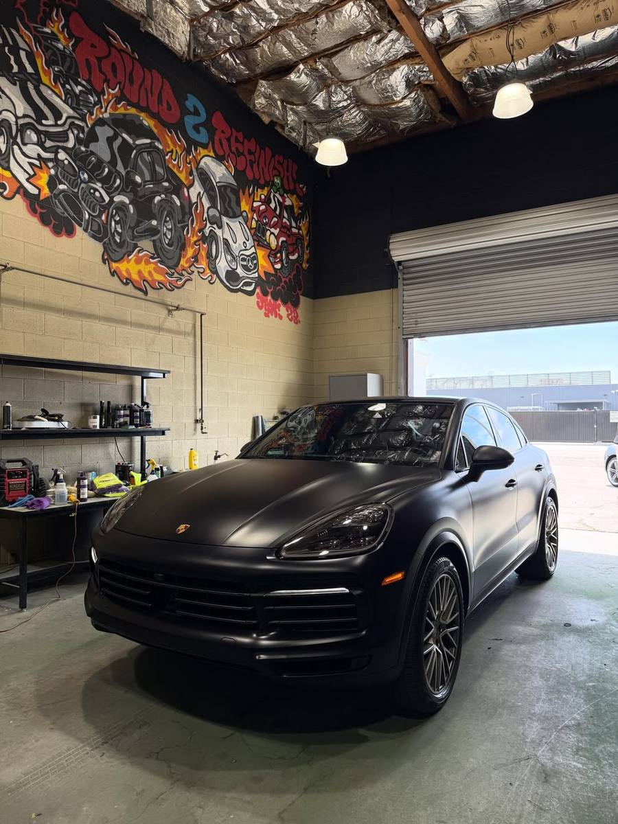 Porsche Cayenne Coupe after a Peel Clear color change, the satin black running unbroken over the hood, bumper, fenders and doors and around the headlights with no seams or cut lines anywhere; parked under the flame mural in the shop
