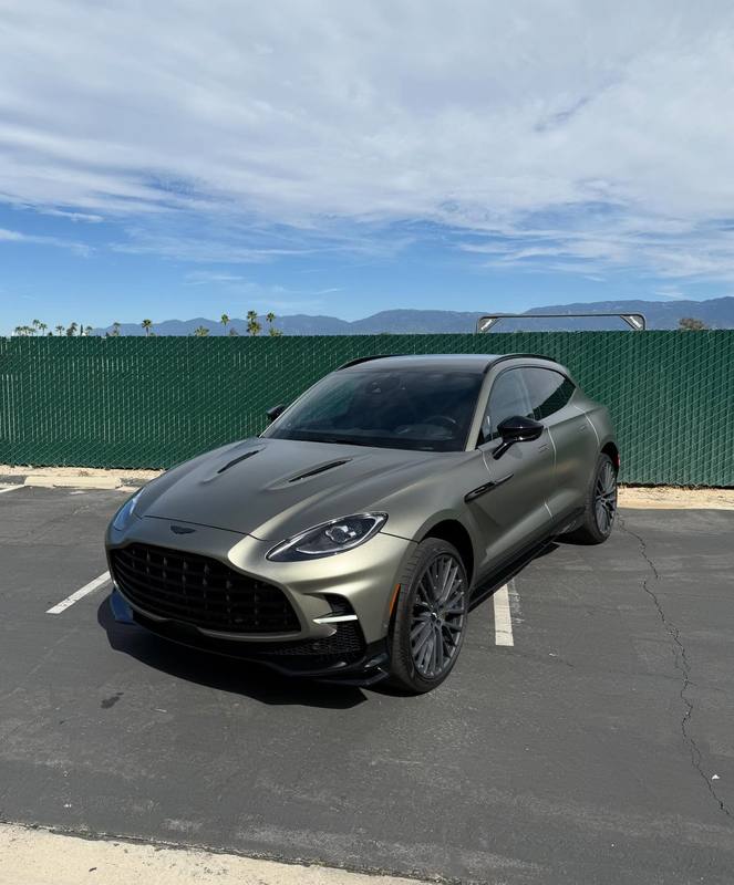 Aston Martin DBX707 in satin olive green, the matte finish flattening the light across the hood vents and the shoulder line so the body shows as shape rather than reflection; dark wheels, black window trim, parked outside the shop