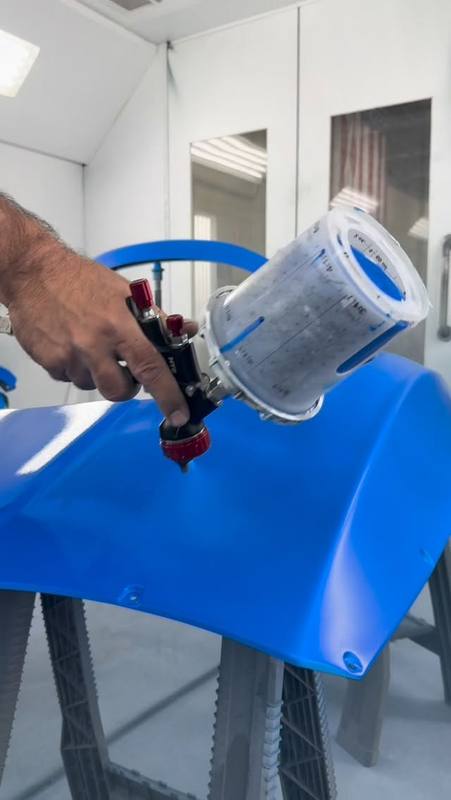 A technician spraying blue Peel Clear from an HVLP gun onto a panel resting on stands in the booth, the coat already laid down wet and even across the curve of the panel