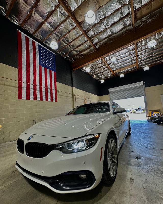 White BMW 4 Series coupe parked inside the shop under the American flag, the hood, bumper and front fender reading as one even white with the flag's stripes reflected faintly across them