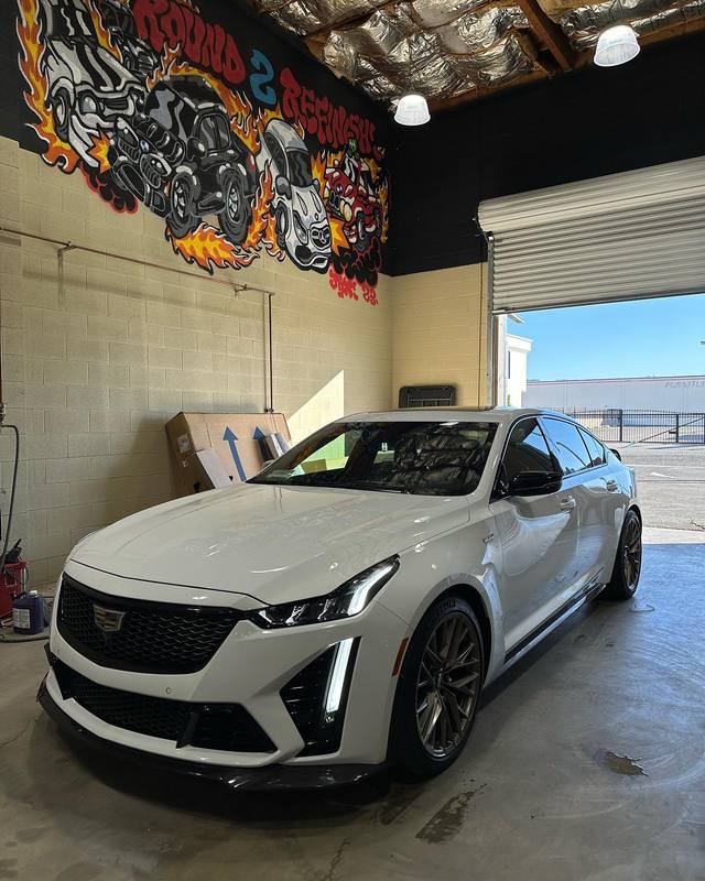 Cadillac CT5-V Blackwing in off-white with dark glass, a black grille, splitter and graphite wheels, parked just inside the open roll-up door with the flame mural on the wall above it