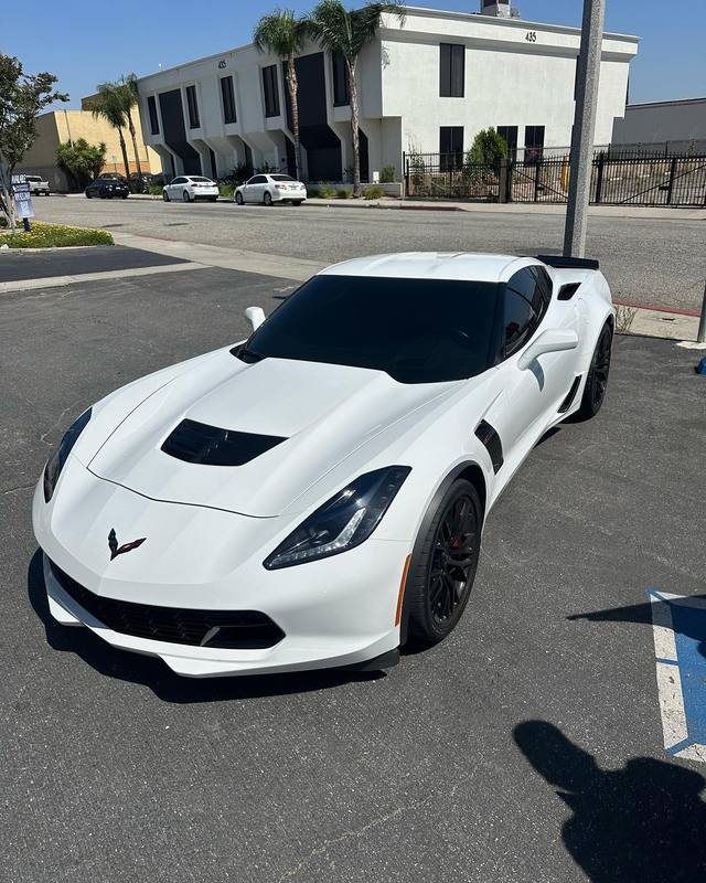 Chevrolet Corvette C7 Z06 in gloss white with a black roof panel, dark glass and black wheels, the white hood and fenders holding a hard edged reflection of the midday sun; parked in the lot outside the shop