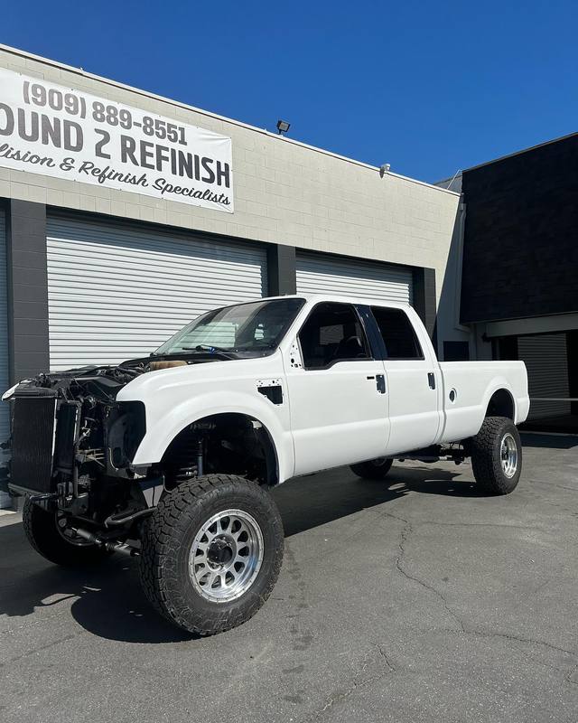 Lifted white Ford F-250 outside the shop with the whole front clip off for collision repair, the grille, bumper, hood and front fender removed and the radiator support, suspension and inner fender left open to view