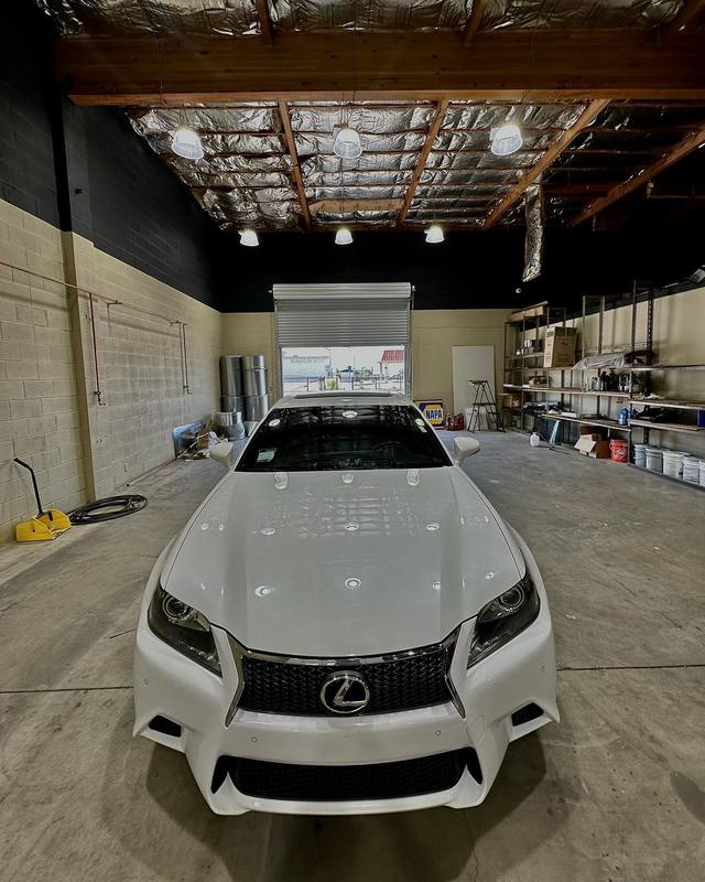 White Lexus GS F Sport shot head-on inside the shop, the row of ceiling lights running evenly down the hood and the hood, fenders and front bumper all matching as one white