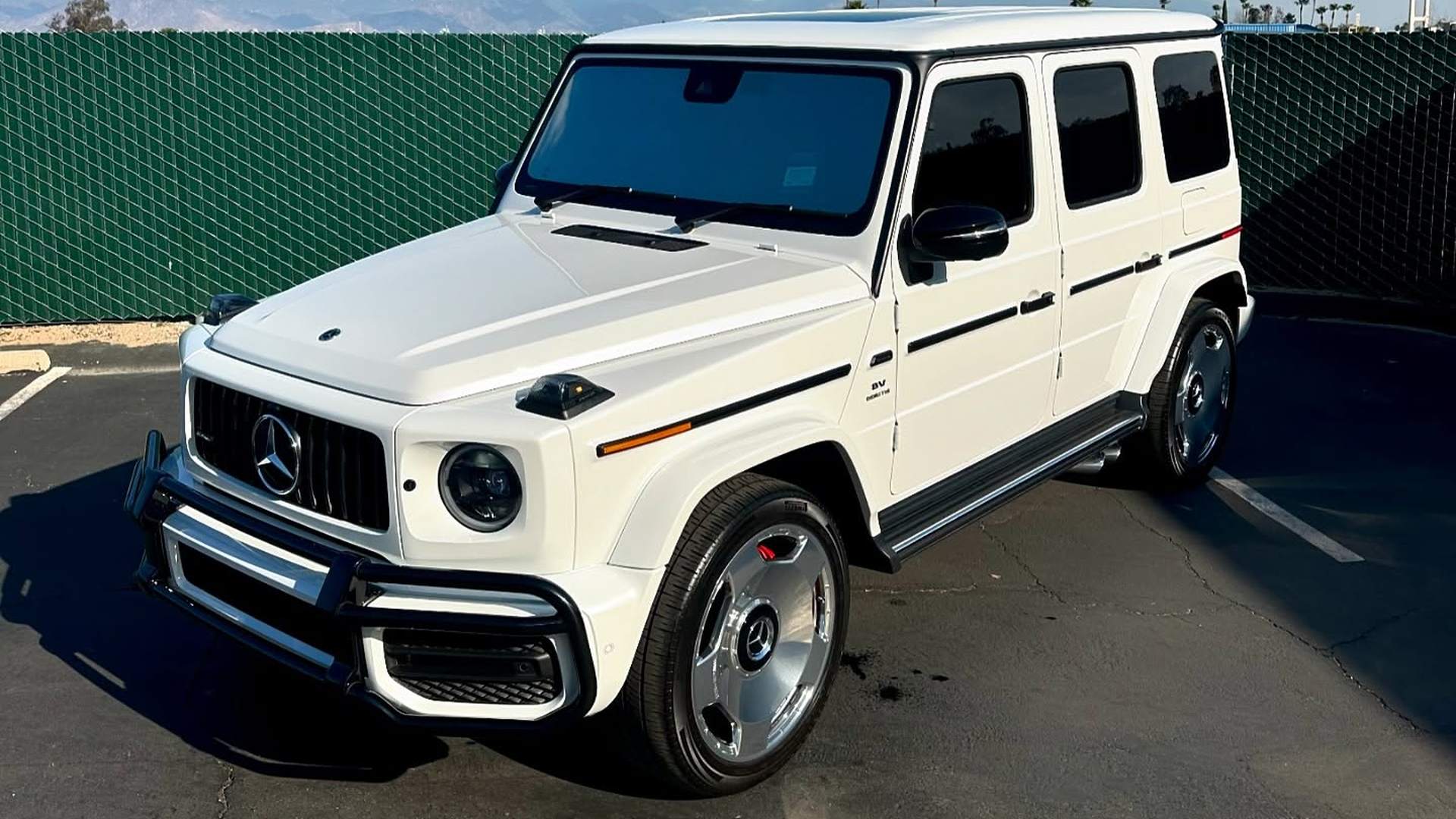 Mercedes-AMG G63 in gloss white with dark glass and black window frames and side moldings, the chrome grille, brush guard and wheels left bright against it, parked in the lot with the mountains behind