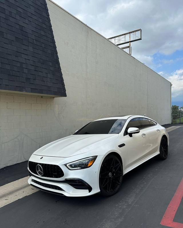Mercedes-AMG GT 53 in gloss white with gloss black wheels and dark glass, parked along the block wall outside the shop, the white body reading flat and even end to end under an overcast sky