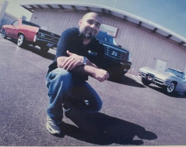 An older photograph of Armen Hovannesian crouching on a shop lot, with a red muscle car, a dark classic pickup and a white Corvette parked behind him