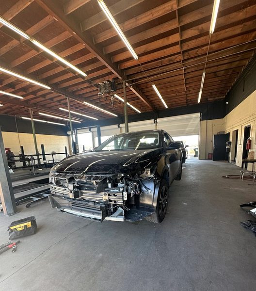 A black SUV in the repair bay with its front bumper cover, grille and headlights taken off, leaving the bumper reinforcement bar, radiator support and wiring exposed, with a frame rack along the wall behind it