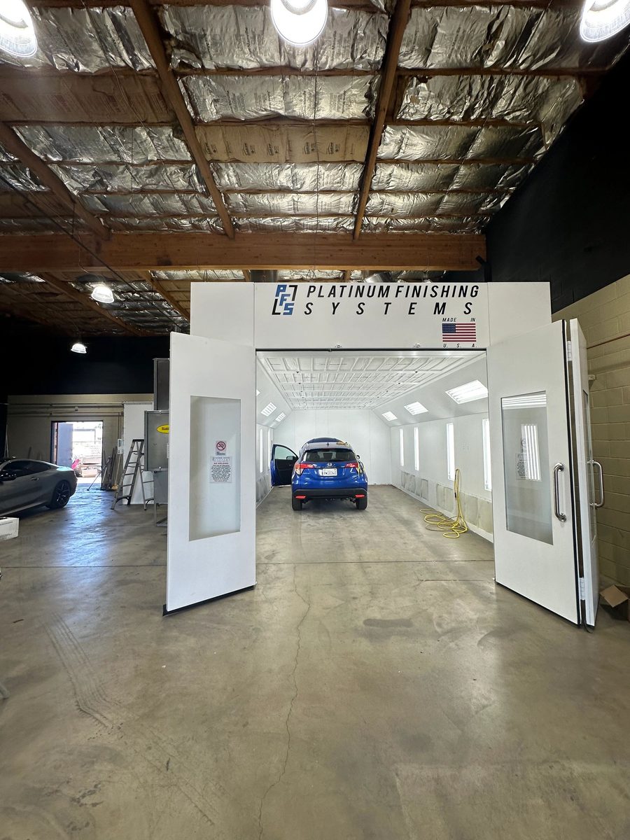 The Platinum Finishing Systems downdraft booth with both doors swung open, a blue Honda HR-V parked nose-in on the booth floor under the white ceiling lights and a yellow air line coiled beside it