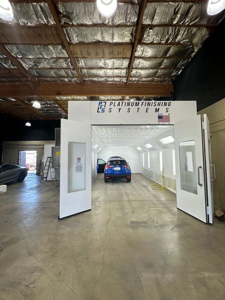The Platinum Finishing Systems downdraft booth with both doors swung open, a blue Honda HR-V parked nose-in on the booth floor under the white ceiling lights and a yellow air line coiled beside it