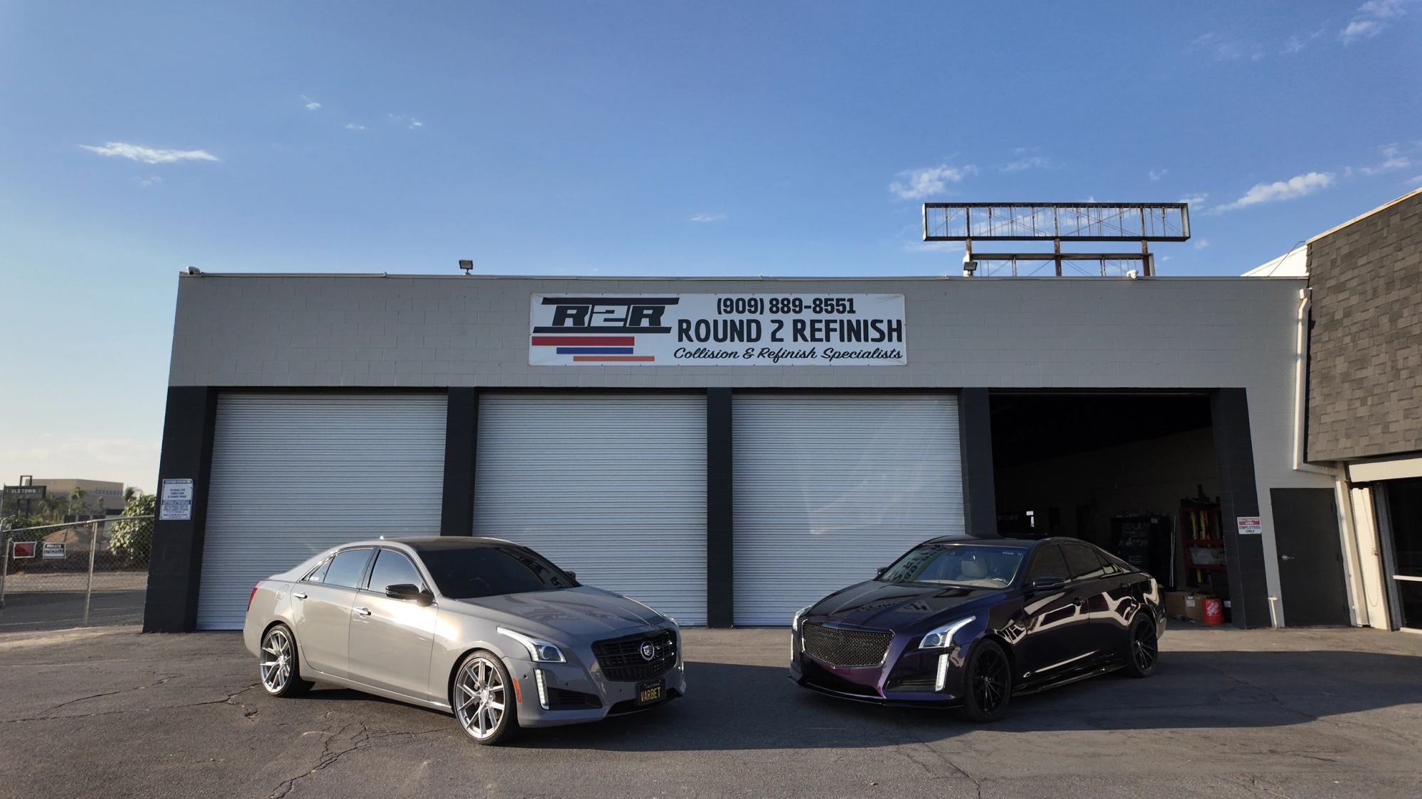 The Round 2 Refinish building on Orange Show Lane, three white roll-up doors under the R2R banner, with a silver Cadillac CTS parked nose-out on the left and one on the right whose paint reads purple in the sun and near black in the shade